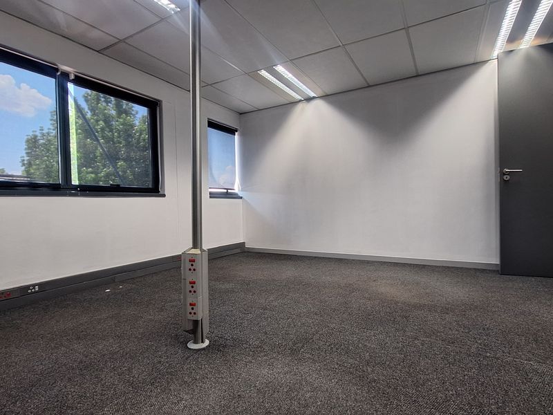 A clean, empty office space featuring carpeted flooring, a central power pole, recessed ceiling lighting, and large windows providing natural light.