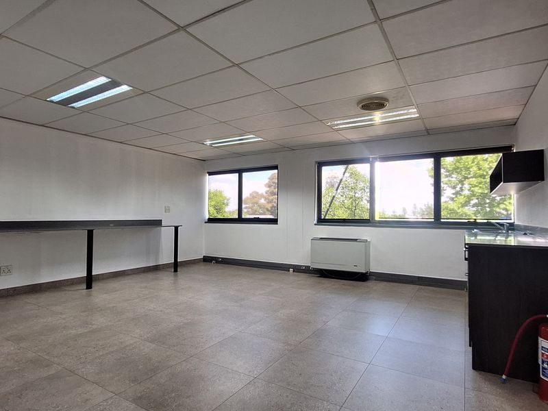 A bright, open-plan office space featuring tiled flooring, a suspended ceiling with recessed lighting, a built-in desk, and a compact kitchenette area.