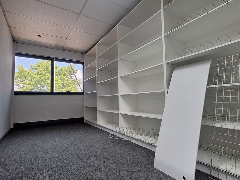 An office space featuring extensive built-in white shelving, grey carpet flooring, and a suspended ceiling with a window providing natural light.