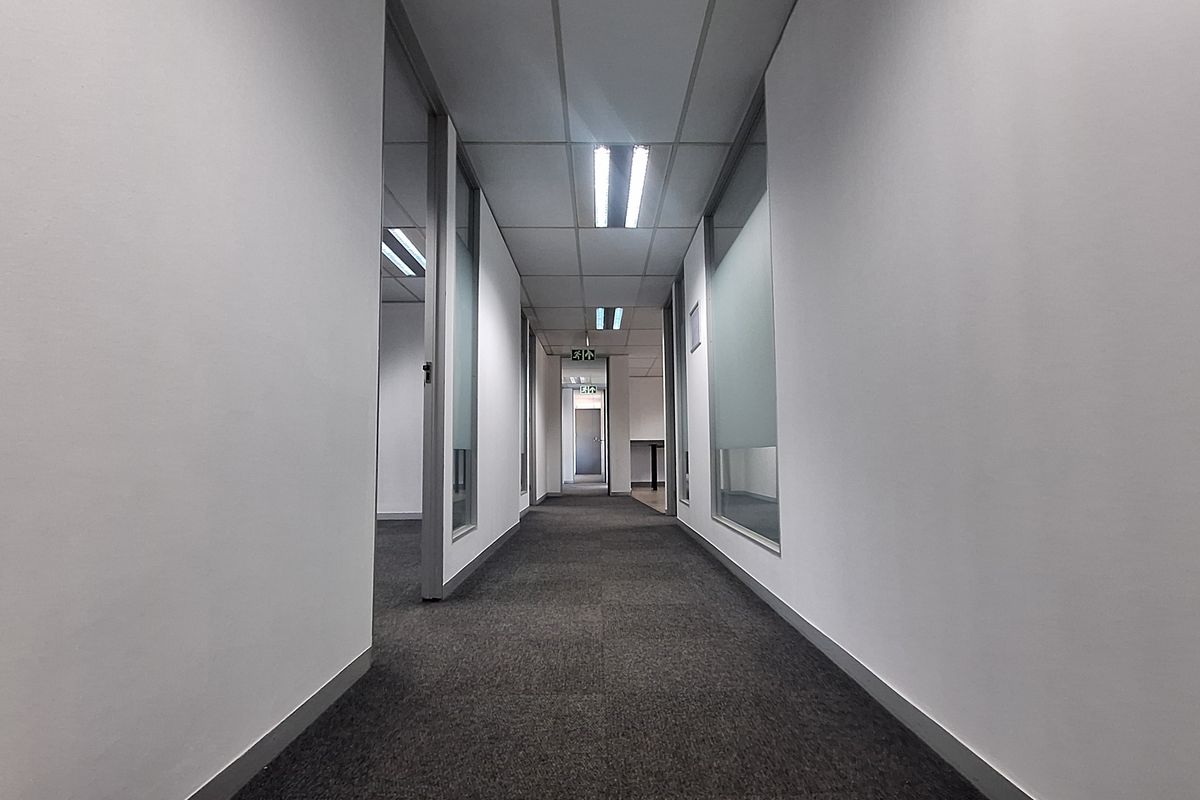 A clean, modern office hallway featuring glass partition walls, carpeted flooring, and a suspended ceiling with recessed lighting.