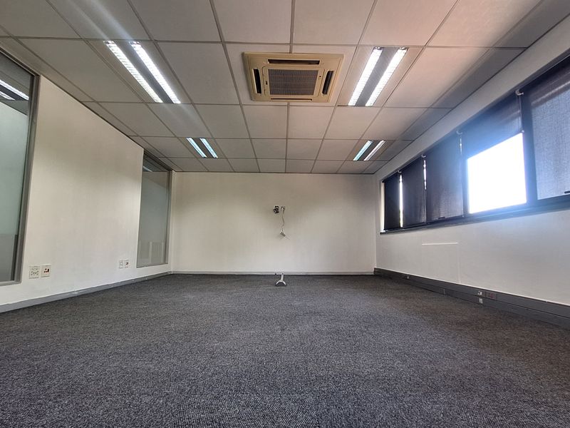 A spacious, empty office featuring a suspended ceiling with recessed lighting, integrated cassette air conditioning, carpeted flooring, and large windows with glass partitions.