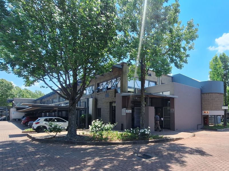 Front exterior view of a modern office building featuring a paved driveway, covered parking bays, and well-maintained landscaped garden areas.