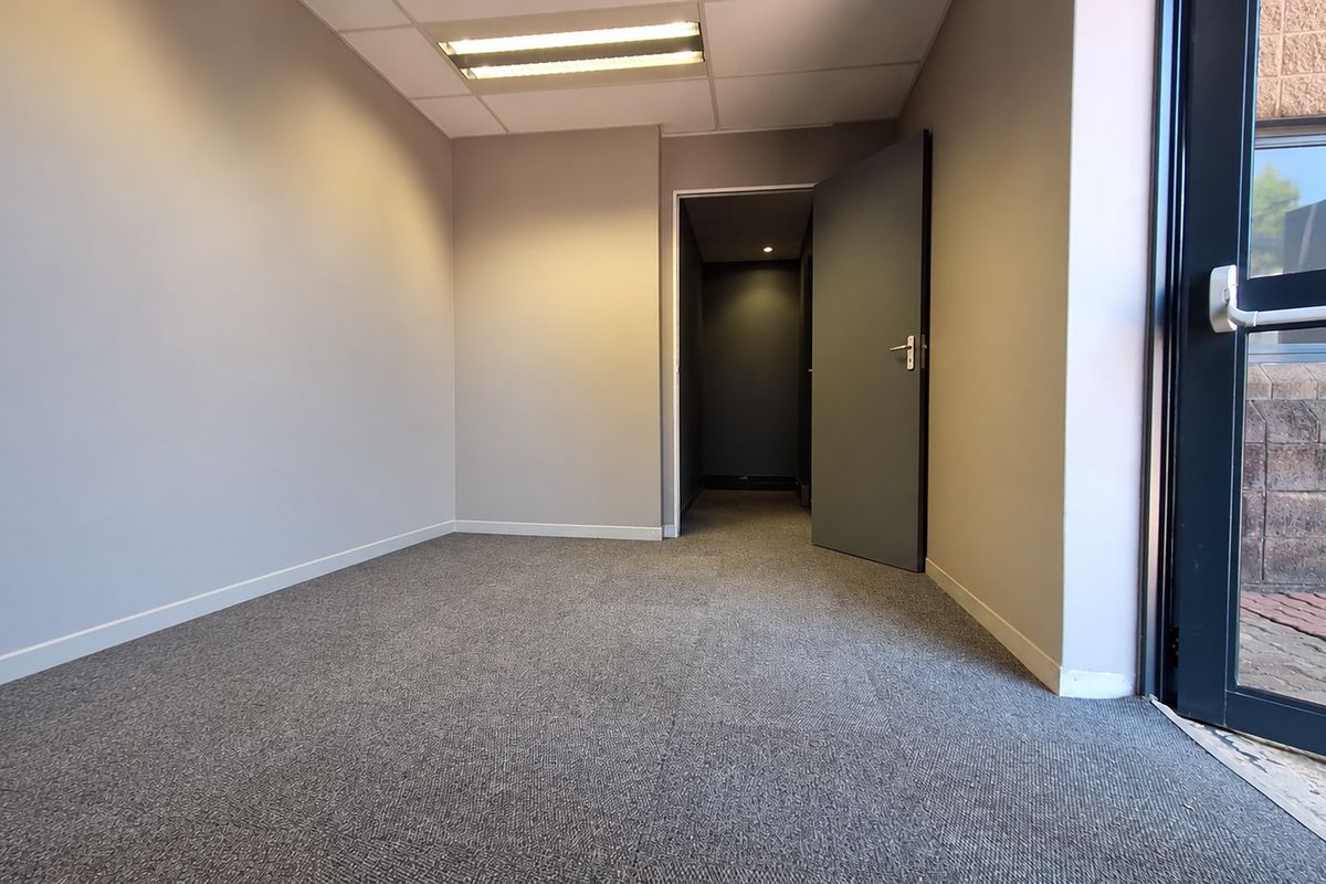 A clean, empty office space featuring neutral grey walls, carpeted flooring, recessed ceiling lighting, and direct access to an exterior entrance.