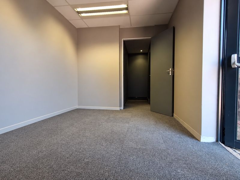 A clean, empty office space featuring neutral grey walls, carpeted flooring, recessed ceiling lighting, and direct access to an exterior entrance.