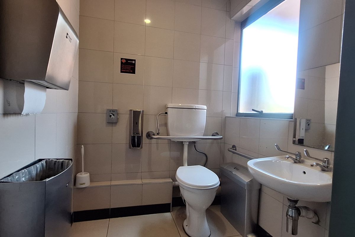 A functional, tiled accessible bathroom featuring a toilet with grab rails, a wall-mounted washbasin, and stainless steel utility dispensers.
