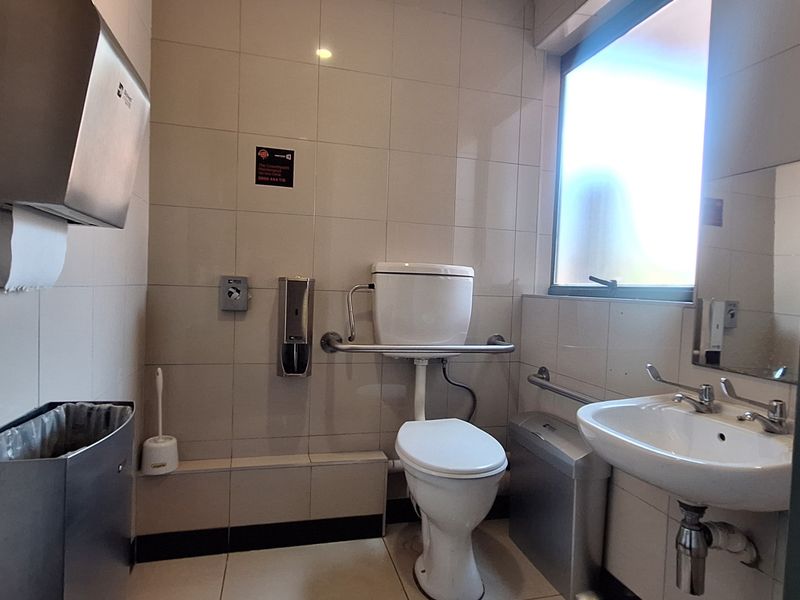 A functional, tiled accessible bathroom featuring a toilet with grab rails, a wall-mounted washbasin, and stainless steel utility dispensers.