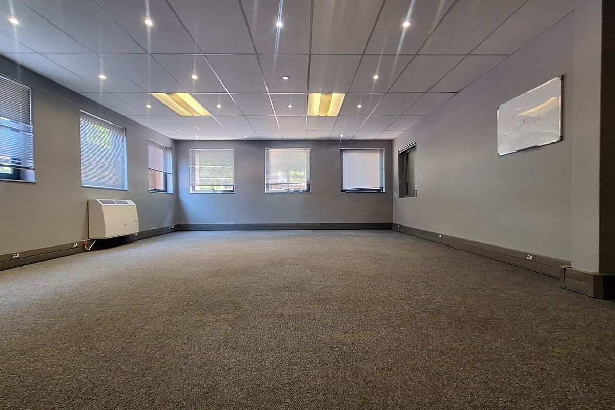 A spacious, empty office area featuring neutral grey walls, carpeted flooring, a suspended ceiling with recessed lighting, and a wall-mounted air conditioning unit.
