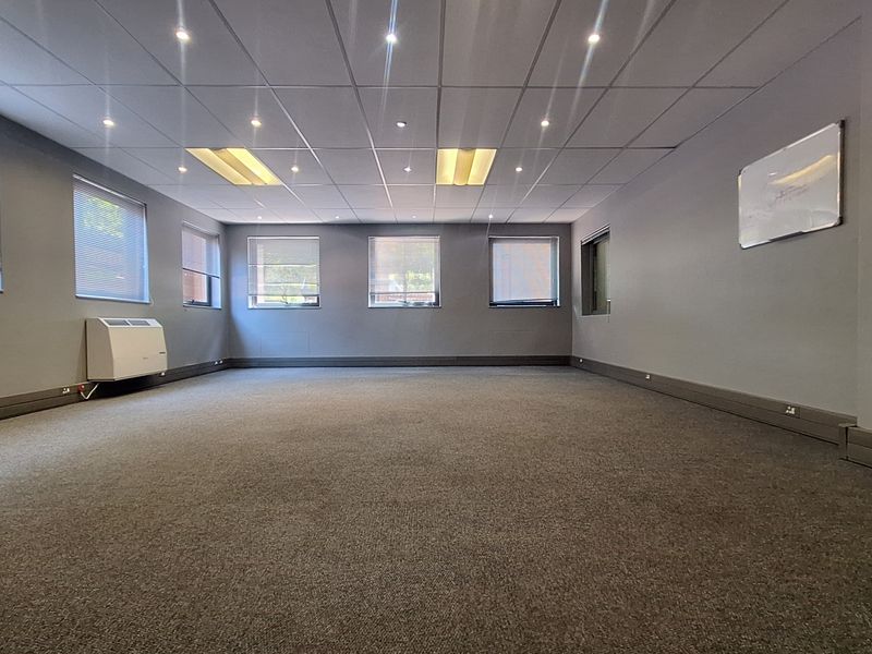 A spacious, empty office area featuring neutral grey walls, carpeted flooring, a suspended ceiling with recessed lighting, and a wall-mounted air conditioning unit.