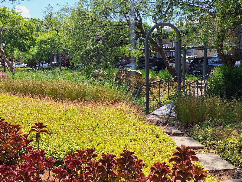 A well-maintained landscaped garden featuring a metal footbridge, paved pathways, and mature trees, providing a pleasant outdoor space for the office property.