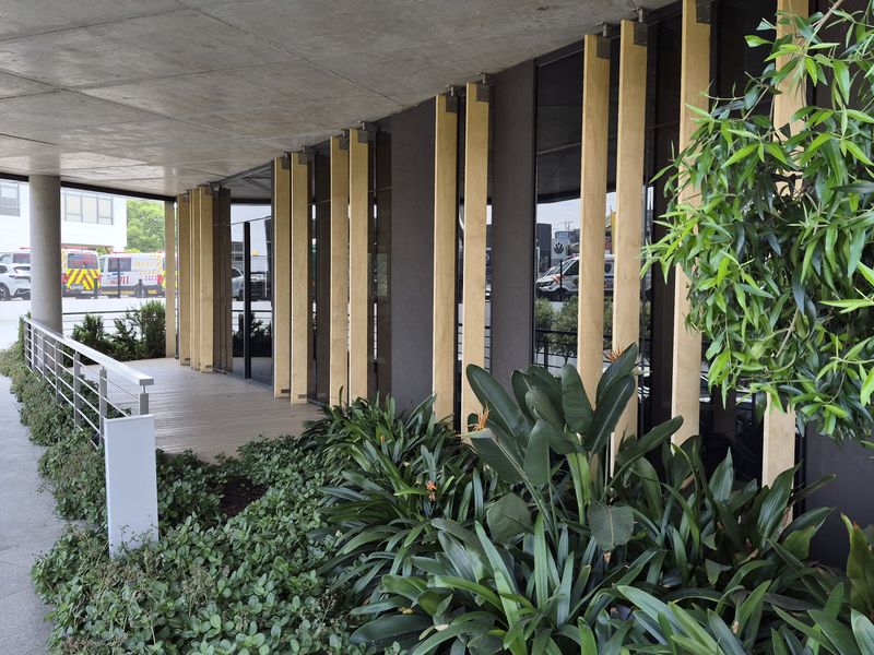 A modern covered patio area featuring a concrete ceiling, timber facade detailing, and glass walling, overlooking a landscaped garden and parking area.