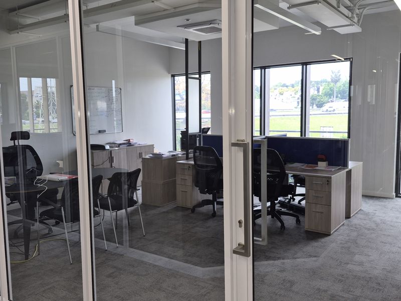 Modern office space featuring glass partition walls, carpeted flooring, and large windows providing ample natural light and views.