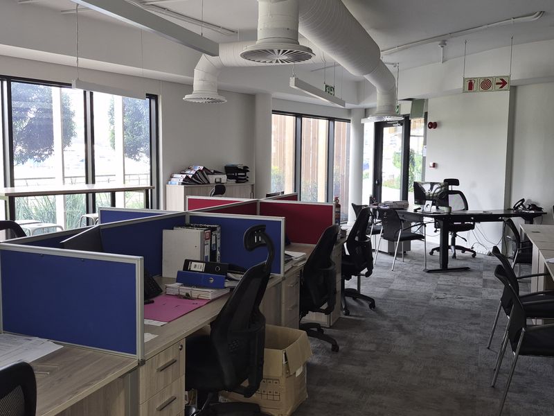 Modern open-plan office space featuring exposed industrial ducting, large windows providing natural light, and carpeted flooring in a professional business environment.