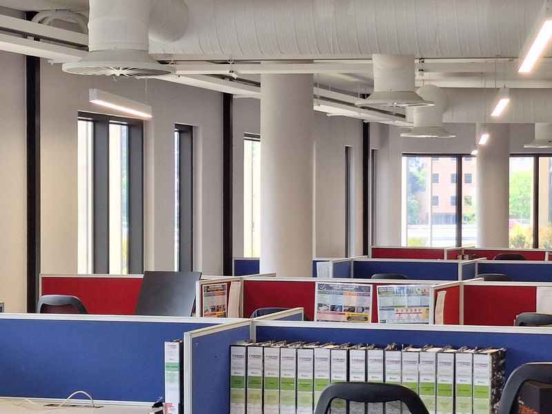 Open-plan office space featuring exposed industrial ductwork, modern linear lighting, structural columns, and integrated cubicle workstations with large windows providing natural light.
