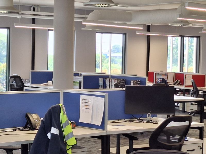 Modern open-plan office space featuring exposed industrial ductwork, suspended linear lighting, and functional cubicle workstations with integrated partitions.