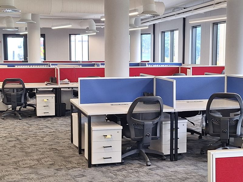 Modern open-plan office space featuring exposed industrial-style ceiling ducting, linear lighting, and integrated desk workstations with privacy partitions.