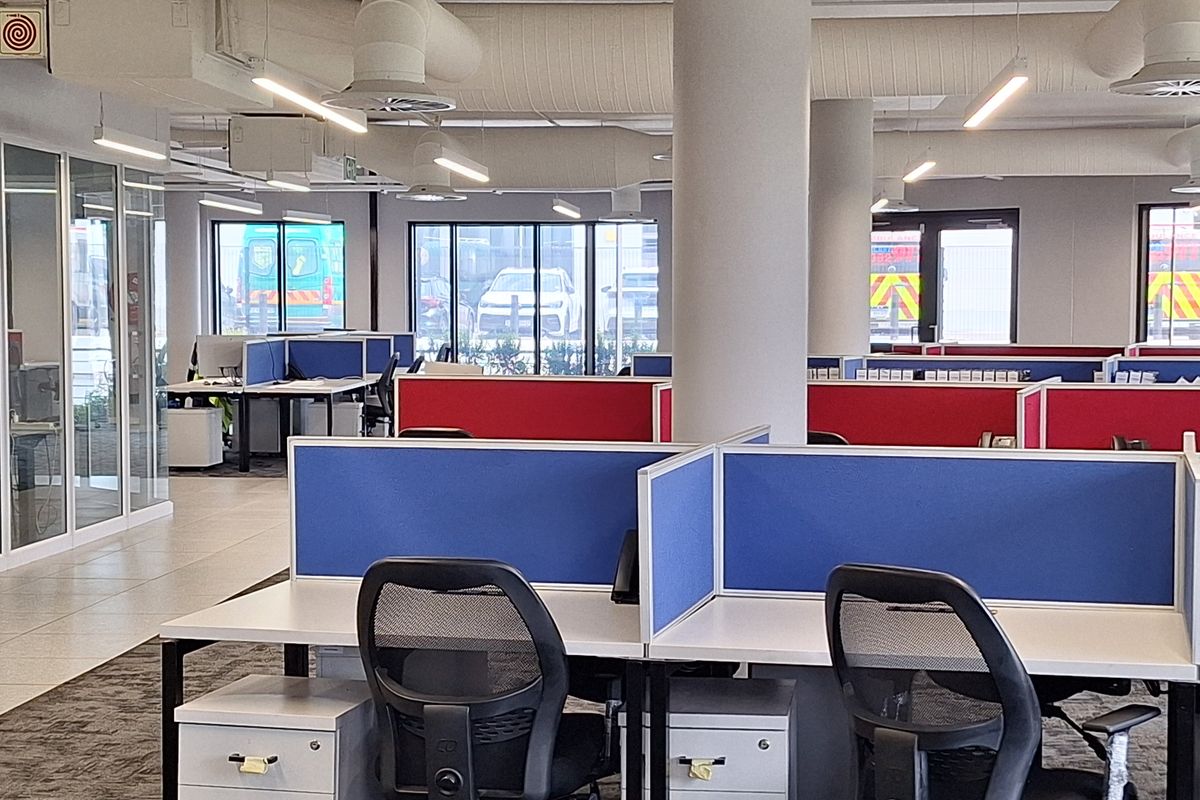 Modern open-plan office space featuring exposed industrial ductwork, structural columns, glass partitioning, and integrated desk workstations with privacy dividers.