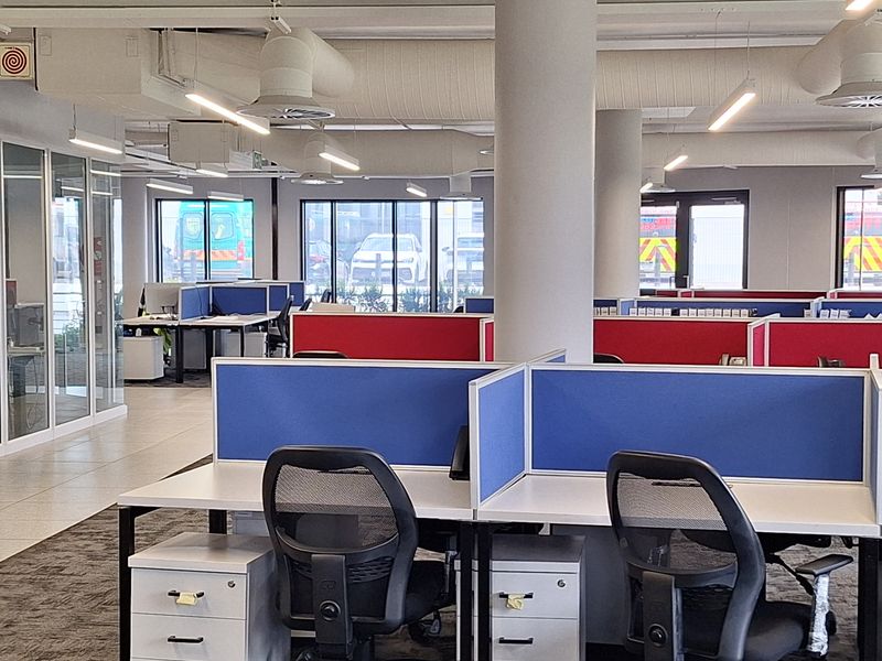 Modern open-plan office space featuring exposed industrial ductwork, structural columns, glass partitioning, and integrated desk workstations with privacy dividers.