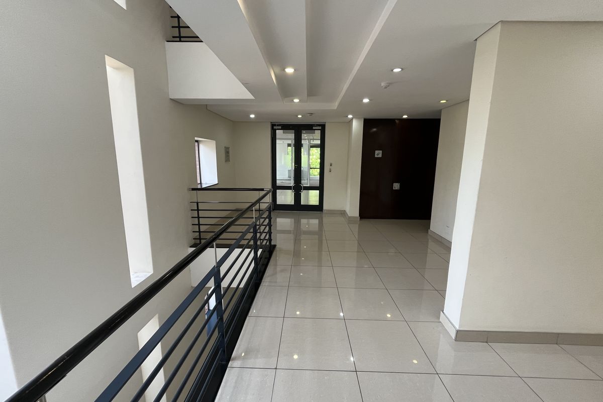 A bright and modern office hallway featuring polished tiled flooring, recessed ceiling lighting, and a sleek metal balustrade overlooking the lower level.