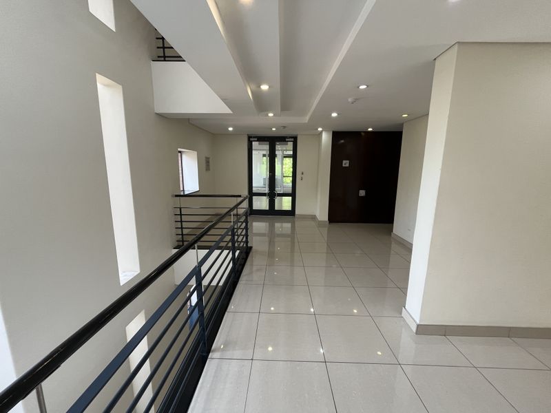 A bright and modern office hallway featuring polished tiled flooring, recessed ceiling lighting, and a sleek metal balustrade overlooking the lower level.
