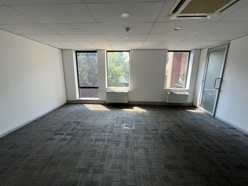 A bright, empty office space featuring carpeted flooring, a suspended ceiling with recessed lighting, and integrated air conditioning units.