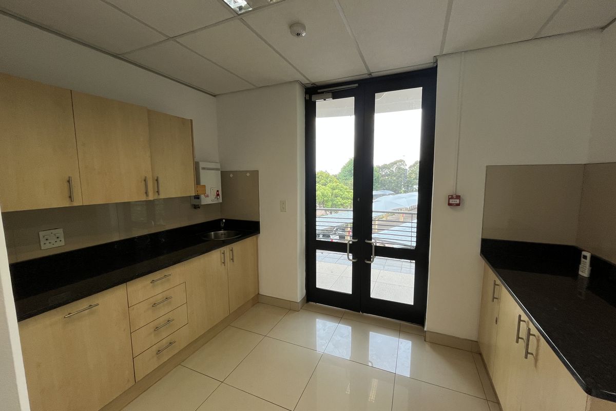 A clean, functional office kitchenette featuring light wood cabinetry, dark granite countertops, tiled flooring, and direct access to an outdoor balcony area.