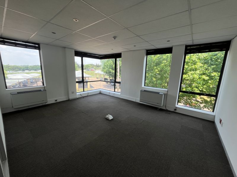 A bright, empty corner office space featuring carpet tiles, a suspended ceiling, and large windows providing ample natural light and exterior views.