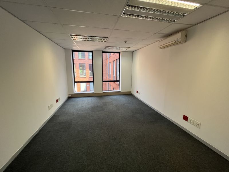 A clean, empty office space featuring carpeted flooring, a suspended ceiling with recessed lighting, and a wall-mounted air conditioning unit.