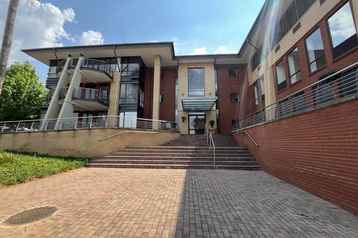 Front elevation of a multi-story office building featuring a brick facade, glass entrance canopy, and paved approach with stainless steel railings.