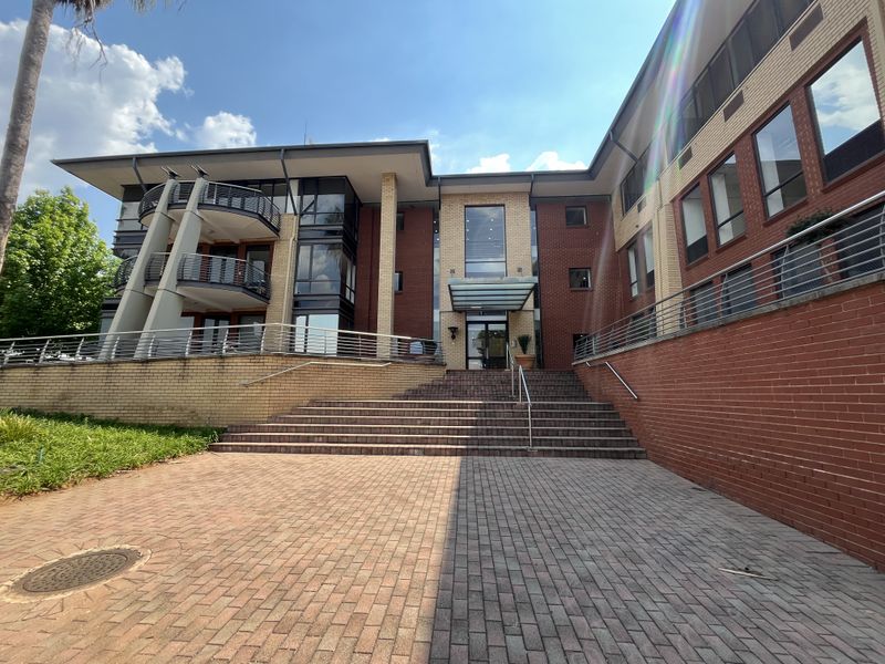 Front elevation of a multi-story office building featuring a brick facade, glass entrance canopy, and paved approach with stainless steel railings.