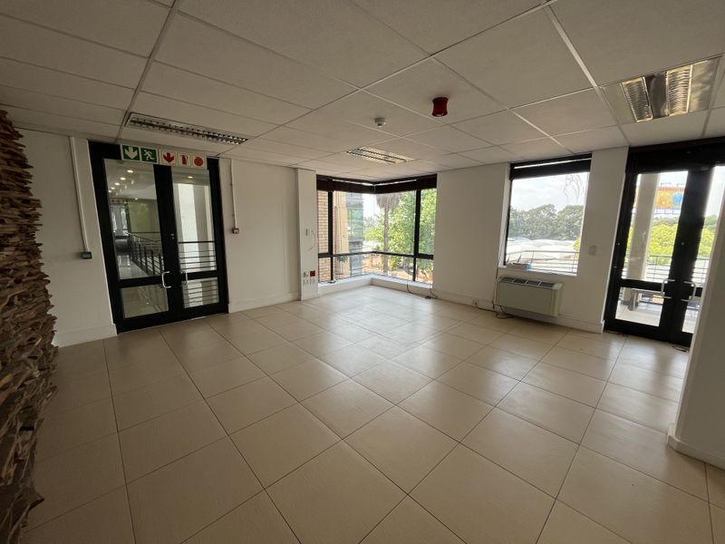 A bright, open-plan office space featuring tiled flooring, a suspended ceiling with recessed lighting, and large windows providing ample natural light.