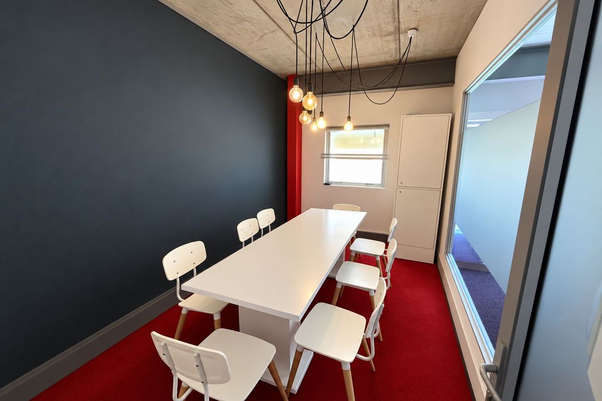 Modern office meeting room featuring an exposed concrete ceiling, vibrant red carpeting, a feature dark wall, and a glass partition for natural light.