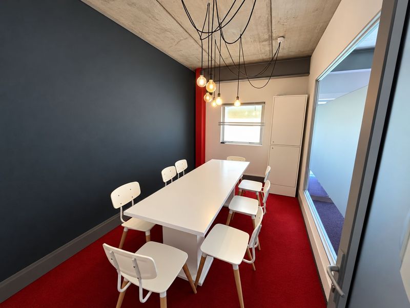 Modern office meeting room featuring an exposed concrete ceiling, vibrant red carpeting, a feature dark wall, and a glass partition for natural light.