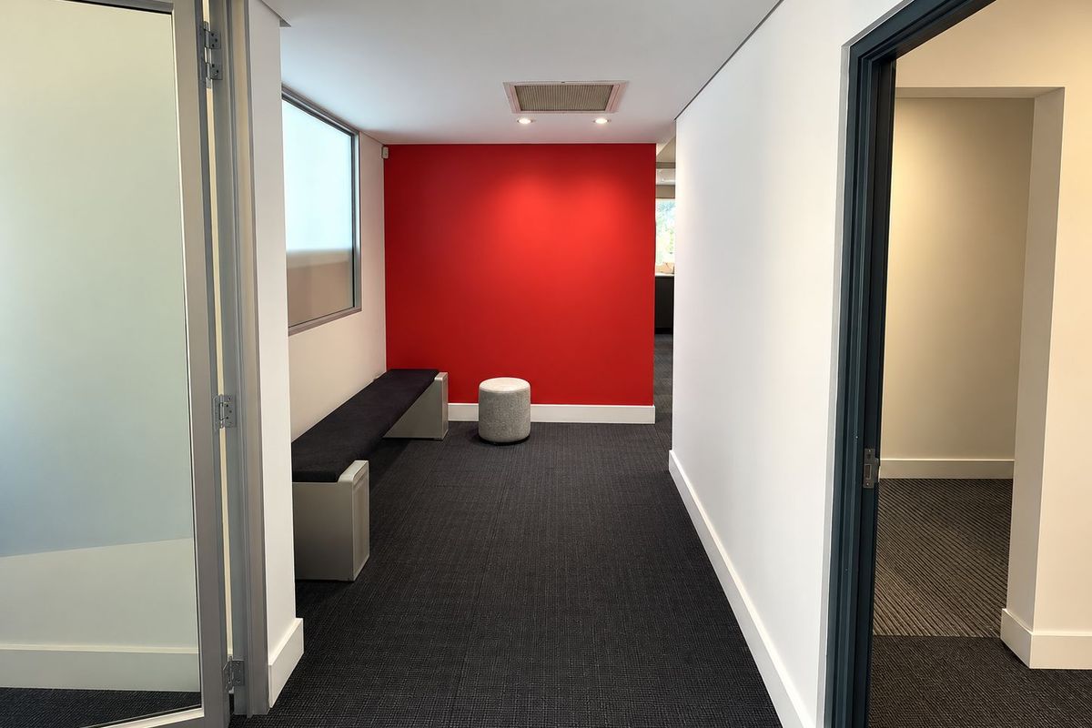 A modern office corridor featuring a bold red accent wall, recessed ceiling lighting, and professional carpet flooring with access to adjacent office spaces.