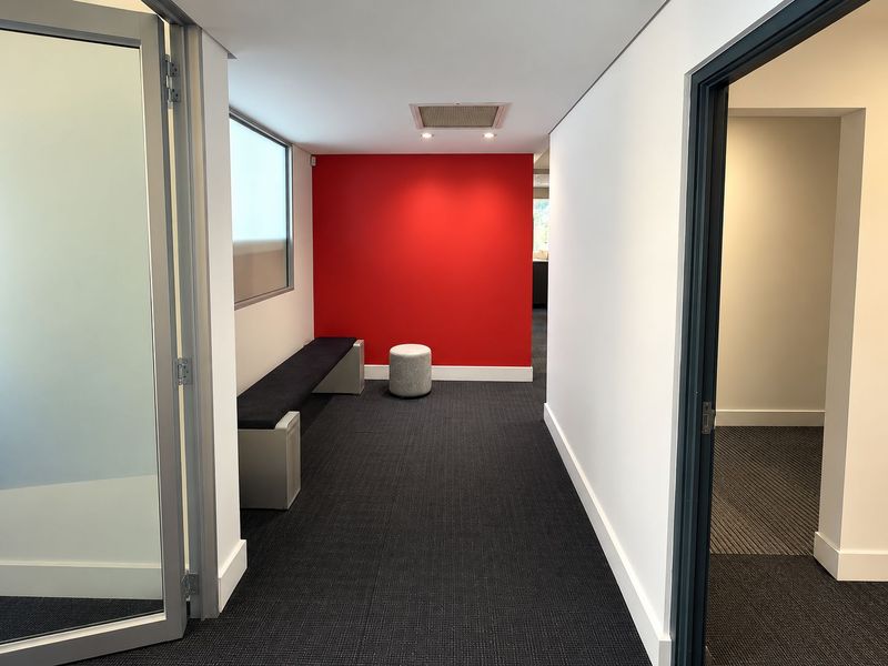 A modern office corridor featuring a bold red accent wall, recessed ceiling lighting, and professional carpet flooring with access to adjacent office spaces.