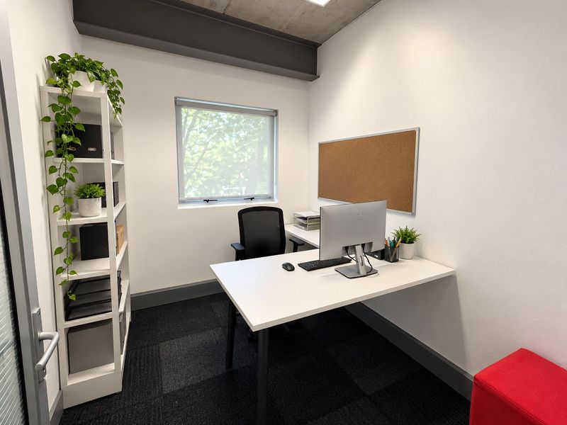 A modern, well-maintained office space featuring an exposed concrete ceiling, steel beam detail, dark carpet tiles, and a functional window with a roller blind.