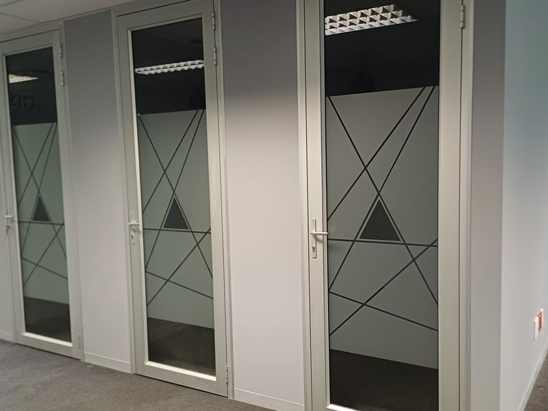 A modern office hallway featuring multiple aluminium-framed glass doors with geometric frosted privacy film, suspended ceilings, and neutral carpeted flooring.