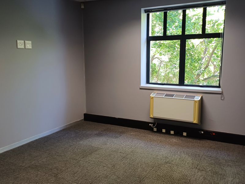 A clean, empty office space featuring carpeted flooring, a large window with natural light, and a wall-mounted air conditioning unit.