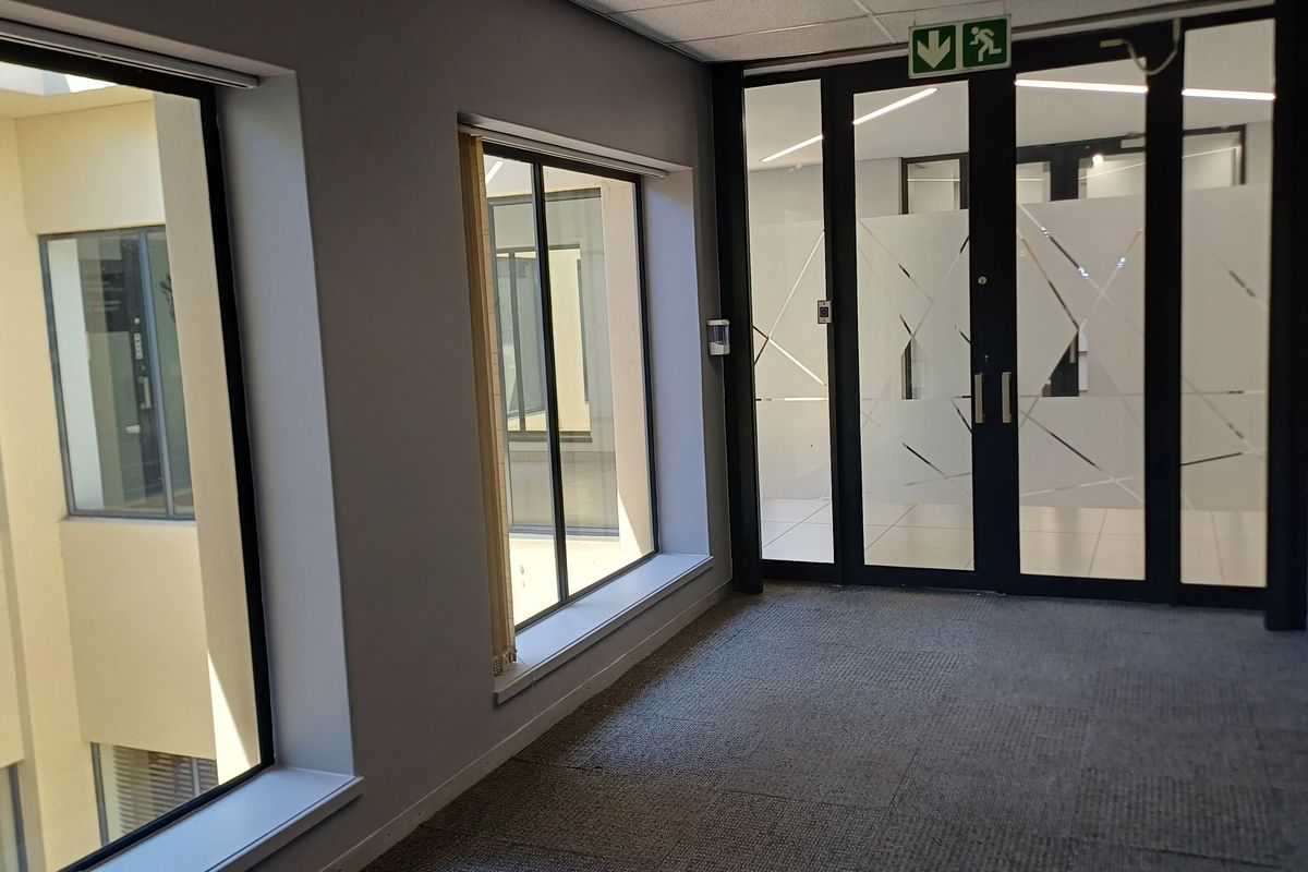 A bright, modern office hallway featuring carpeted flooring, large windows overlooking an internal courtyard, and glass-paneled double doors with emergency exit signage.