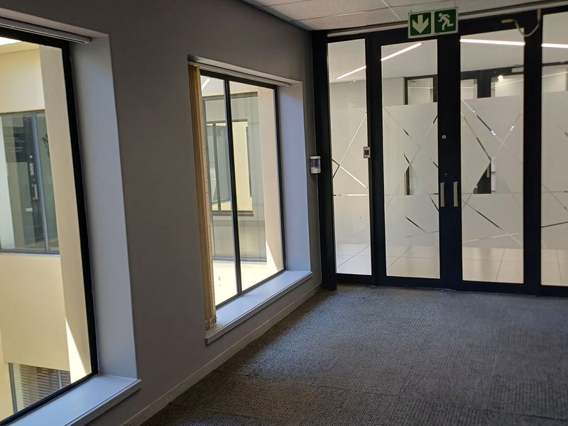 A bright, modern office hallway featuring carpeted flooring, large windows overlooking an internal courtyard, and glass-paneled double doors with emergency exit signage.