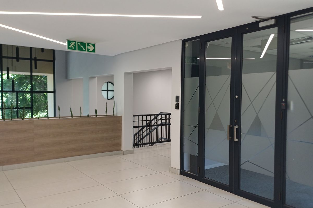 Modern office building entrance lobby featuring tiled floors, linear lighting, glass partition doors, and a staircase leading to upper levels.