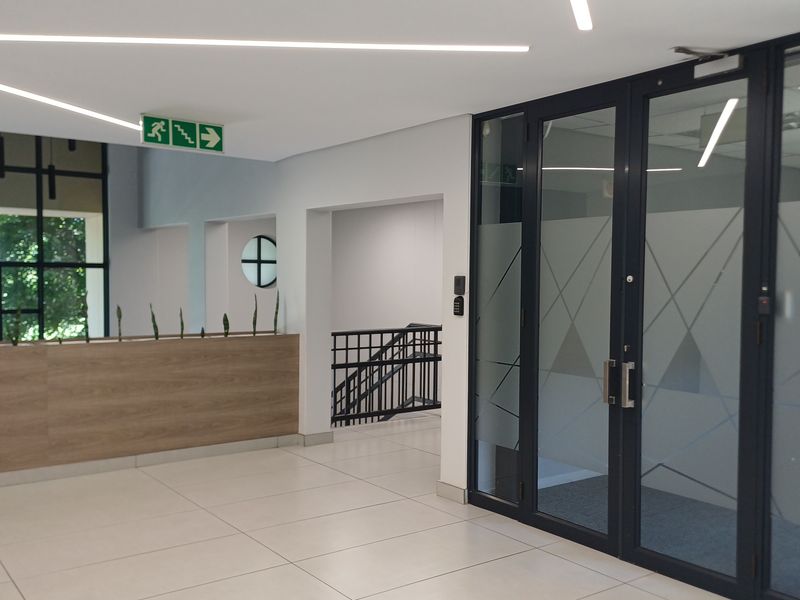 Modern office building entrance lobby featuring tiled floors, linear lighting, glass partition doors, and a staircase leading to upper levels.