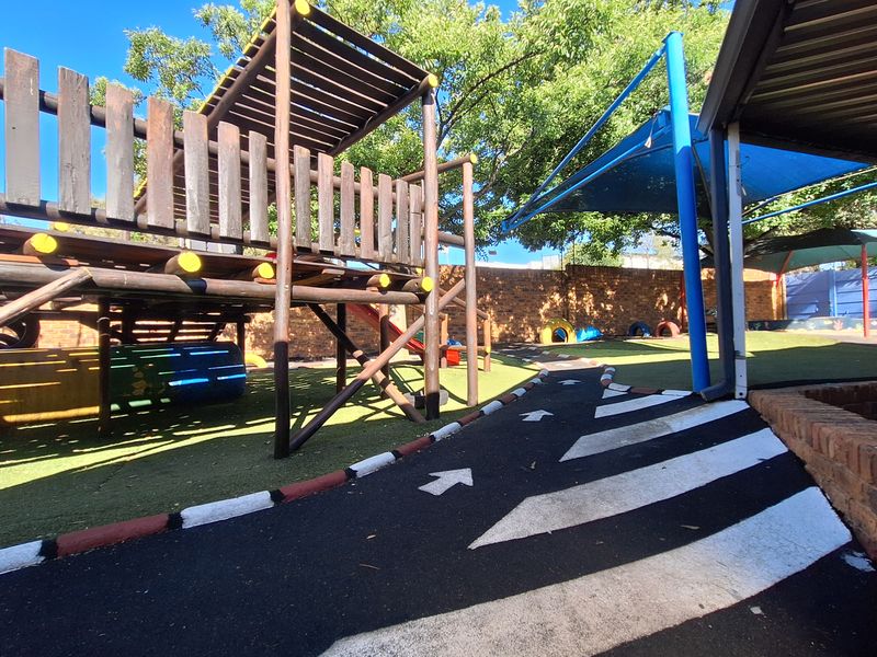 Outdoor commercial play area featuring a wooden climbing structure, artificial turf, shade sails, and a painted asphalt track for children.
