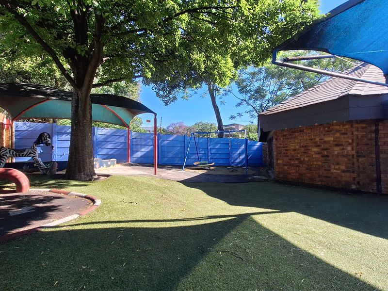 Outdoor play area featuring artificial turf, shade sails, a swing set, and brick boundary walls in a commercial setting.