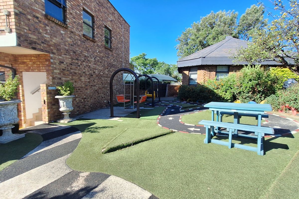 Rear exterior area featuring face brick building facades, artificial turf landscaping, paved pathways, and fixed playground equipment in a commercial setting.