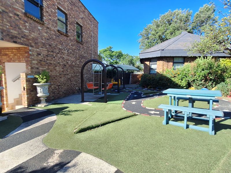 Rear exterior area featuring face brick building facades, artificial turf landscaping, paved pathways, and fixed playground equipment in a commercial setting.