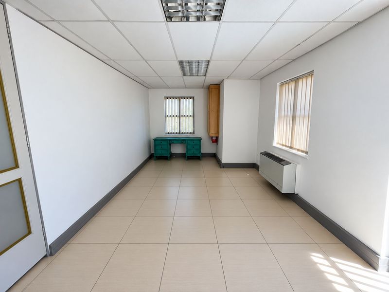 A bright, empty office space featuring tiled flooring, a suspended ceiling with recessed lighting, wall-mounted HVAC unit, and vertical blinds on the windows.