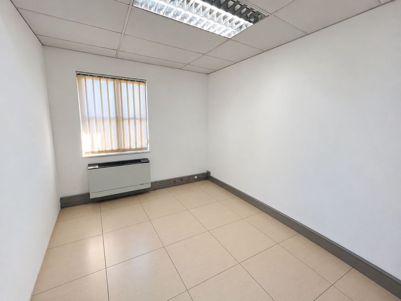 A clean, empty office space featuring tiled flooring, a suspended ceiling with recessed lighting, and a wall-mounted HVAC unit.