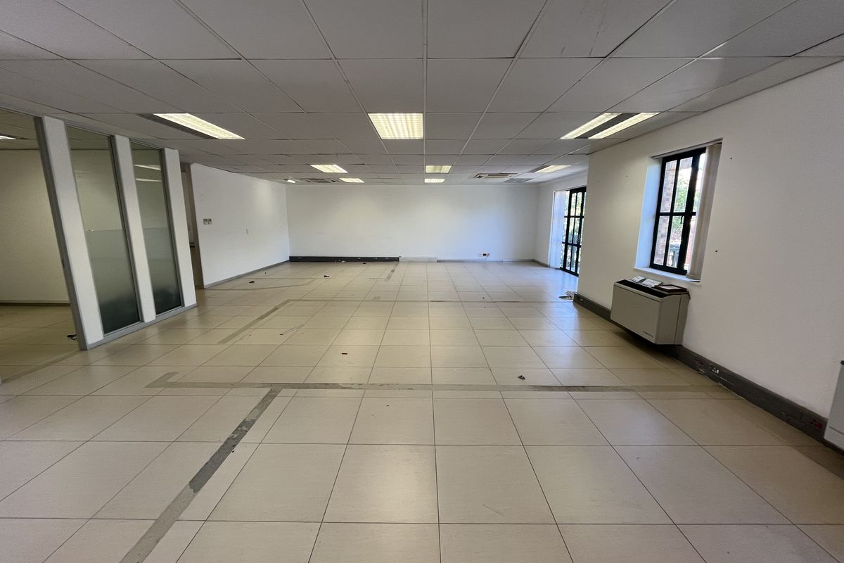 A spacious, empty open-plan office area featuring tiled flooring, a suspended ceiling with recessed lighting, and glass partition walls.