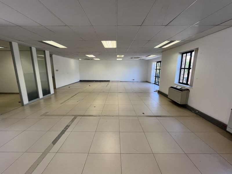 A spacious, empty open-plan office area featuring tiled flooring, a suspended ceiling with recessed lighting, and glass partition walls.