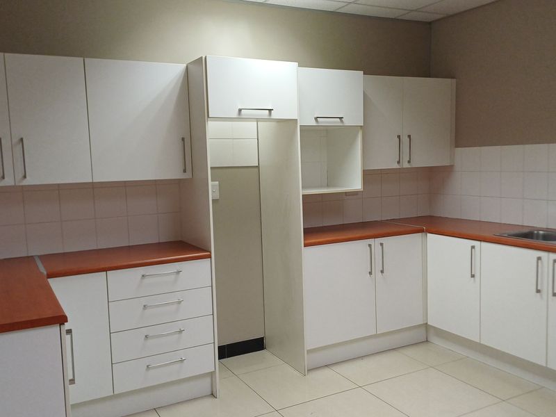 A clean, functional office kitchenette featuring white cabinetry, laminate countertops, a stainless steel sink, and tiled backsplash.